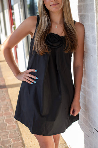 Model wearing a sleeveless black swing dress with a statement rosette neckline and relaxed above-the-knee silhouette.