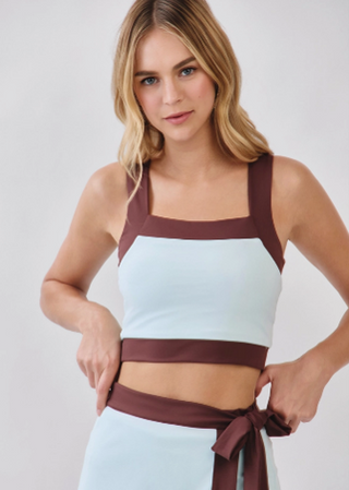 A woman modeling a color-blocked crop top and matching skirt. The top features a light blue body with dark brown straps and a matching brown waistband, while the skirt is light blue with a wide brown waist tie.