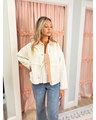 Woman wearing the Amanda cream jacket with orange gingham contrast trim, snap-front closure, and relaxed fit layered over a white top and blue jeans.
