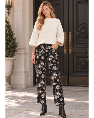 A woman stands outdoors in front of a large dark wooden door wearing a cream wide-sleeve top and black wide-leg pants with a beige floral print, paired with black ankle boots.