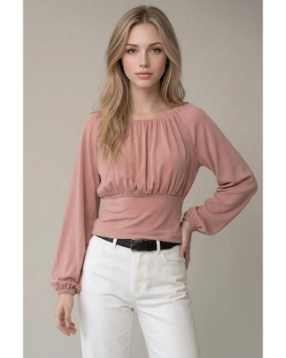 A woman modeling a dusty rose long-sleeve blouse with a gathered, pleated bodice and elasticized cuffs, paired with white pants and a black belt, against a neutral beige background.