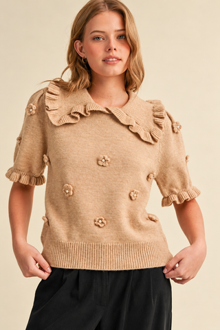 Model wearing a camel beige women's knit sweater with a ruffled collar, short puff sleeves, pearl-centered 3D floral appliqués, and ribbed hem, styled with black pants.