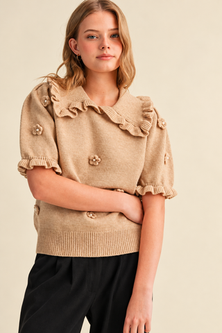 Model wearing a camel beige women's knit sweater with a ruffled collar, short puff sleeves, pearl-centered 3D floral appliqués, and ribbed hem, styled with black pants.