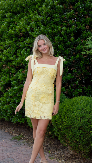 A woman wearing a pale yellow lace mini dress with tie shoulder straps, standing outdoors in front of a large green hedge on a brick pathway.