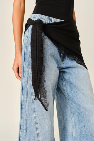 Close-up of a person wearing light-wash wide-leg jeans with a black sheer lace wrap tied around the waist over a black top, against a plain light beige background.
