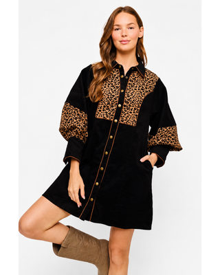 Woman wearing a black button-front mini shirt dress with leopard print chest and sleeve panels, gold-tone buttons, side pockets and tan western boots.