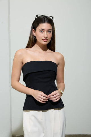 Model wearing a black strapless peplum top with a fitted bodice and flared hem paired with white tailored pants.