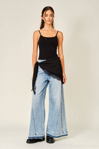 A model wearing a black camisole top, wide-leg light blue denim jeans, and black shoes, standing against a plain light beige studio background.