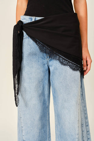 A close-up of a person wearing light blue wide-leg jeans with a black wrap-style top tied at the waist and black lace trim, against a plain light beige background.