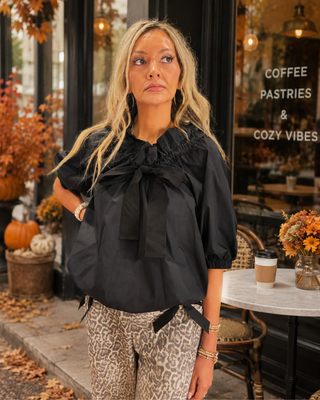 A woman wearing a black puff-sleeve blouse with a large bow at the neckline and leopard-print pants, standing on a sidewalk in front of a cafe storefront with autumn decorations.
