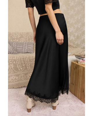 A woman wearing a long black satin maxi skirt with lace trim at the hem, paired with a black lace short-sleeve crop top and beige ankle boots. 