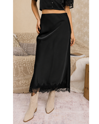Woman wearing a black satin high-waisted midi skirt with delicate black lace trim at the hem, styled with cream pointed-toe ankle boots.