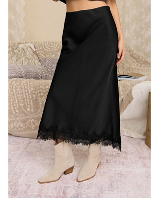 A black satin midi skirt with a delicate lace hem, modeled with cream-colored ankle boots.