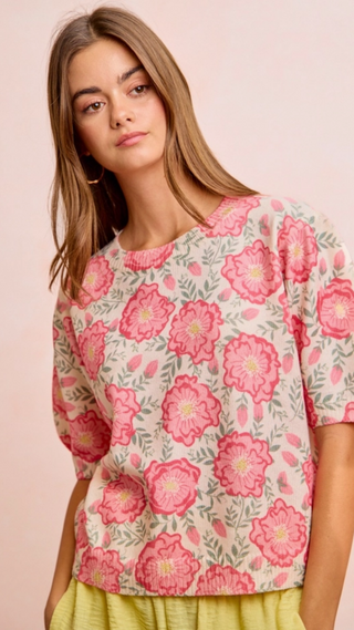 A woman wearing a short-sleeve crewneck top with a pink and green floral pattern on a light beige background, paired with a yellow skirt, set against a solid pale pink studio backdrop.