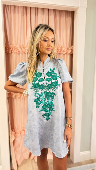 A woman modeling a light blue-gray short-sleeve tunic dress with a green floral lace applique on the front, standing in front of pink curtains.
