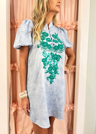 A woman wearing a light blue short-sleeve tunic dress with a green floral embroidered pattern on the front, standing in front of pink curtains.