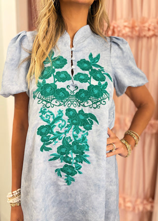 A woman wearing a light blue tunic dress with a green floral embroidered pattern on the front, standing in front of a pink curtain.