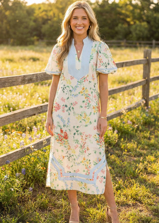 Woman in a floral dress standing in a field with a wooden fence and trees in the background.