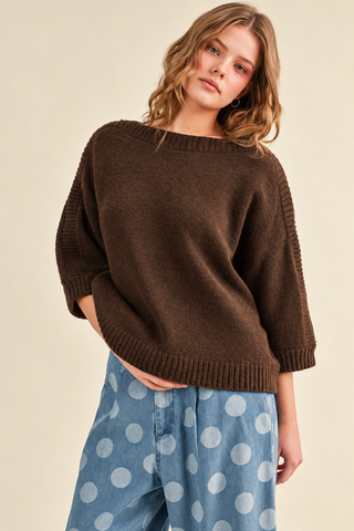 Woman wearing a chocolate brown boat neck knit sweater with three-quarter balloon sleeves, ribbed detailing, and a relaxed fit, paired with light blue polka dot jeans.