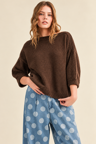 Woman wearing a chocolate brown boat neck knit sweater with three-quarter balloon sleeves, ribbed detailing, and a relaxed fit, paired with light blue polka dot jeans.