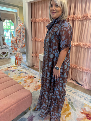 Woman wearing a chocolate brown maxi dress with dusty blue floral print, smocked mock neckline, short flutter sleeves, relaxed fit, and ruffled hem, styled with tan boots.