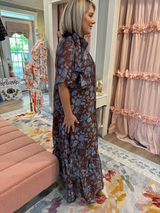 Woman wearing a chocolate brown maxi dress with dusty blue floral print, smocked mock neckline, short flutter sleeves, relaxed fit, and ruffled hem, styled with tan boots.