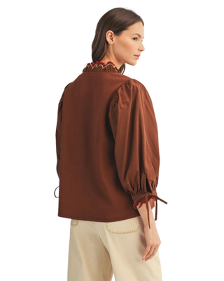 Women’s chocolate brown blouse with red scalloped trim, decorative V-neckline, puff sleeves and tie cuffs, styled with cream pants.