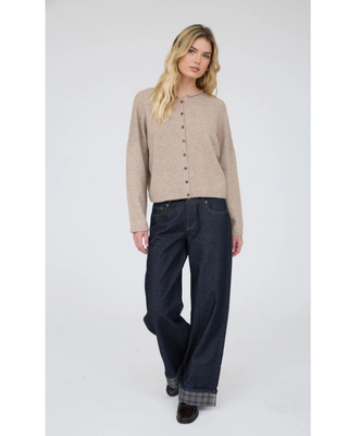 A blonde woman wearing a light beige knit cardigan with a buttoned front and dark blue jeans, photographed against a neutral light gray studio background.