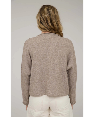 A blonde woman wearing a light beige knit cardigan with a buttoned front and dark blue jeans, photographed against a neutral light gray studio background.