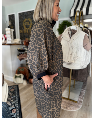 Woman wearing an olive and black leopard print quarter zip mini dress with long sleeves, black trim, side pockets, and tan knee-high boots.