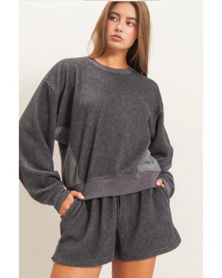 A woman wearing a dark gray heathered lounge set consisting of a long-sleeve sweatshirt and matching shorts, standing against a plain off-white background.