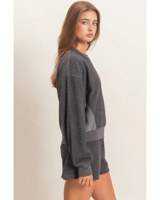 A woman modeling a dark gray washed crewneck sweatshirt and matching shorts against a plain beige studio background.