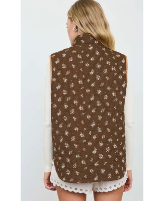 Woman wearing a chocolate brown quilted vest with a delicate floral print, stand-up collar, snap-button and zipper closure, and front patch pockets over a cream ribbed top.