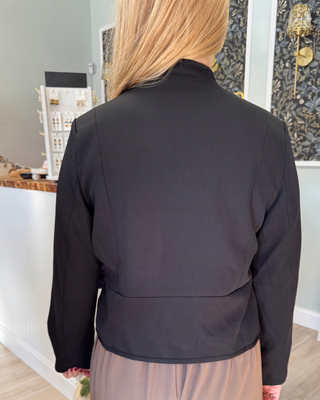A woman viewed from behind wearing a black long-sleeve cropped jacket with structured shoulders and a high mock neck, paired with a beige skirt in a boutique setting.