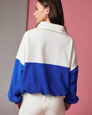 A woman shown from the back wearing a color-block knit sweater with a white upper section, royal blue lower section, and matching blue sleeves, paired with white pants against a split gray and pink background.