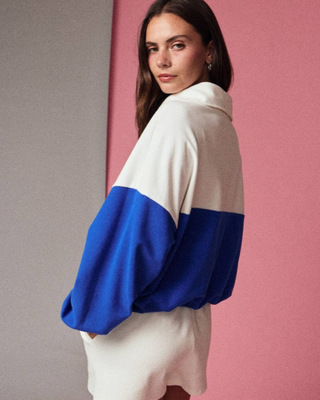 A woman wearing a colorblock quarter zip with a cream upper section and bright blue lower sleeves and hem, posed against a split gray and pink background.