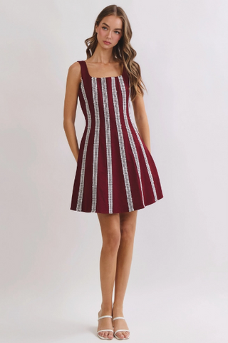 A woman modeling a sleeveless mini dress with vertical burgundy and white textured stripes, standing against a light gray studio background.