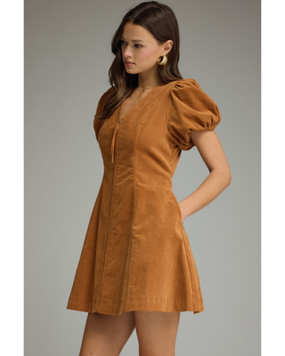 Woman wearing a camel brown corduroy mini dress with short puff sleeves, V-neckline, fitted waist and A-line skirt.
