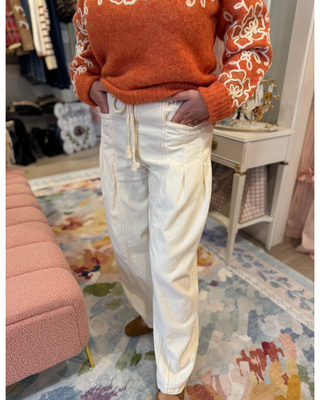 A person wearing cream-colored wide-leg drawstring pants and an orange sweater with white floral embroidery, standing in a styled room with a pink bench, white side table, and a colorful patterned rug.