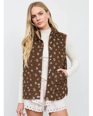 Woman wearing a chocolate brown quilted vest with a delicate floral print, stand-up collar, snap-button and zipper closure, and front patch pockets over a cream ribbed top.