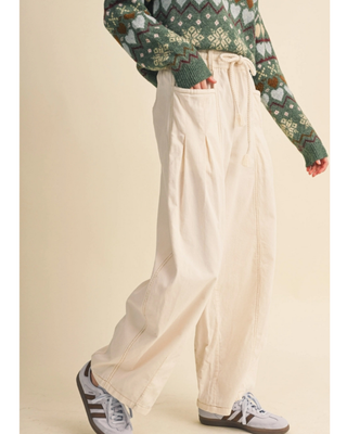 A person wearing cream wide-leg pleated pants with a drawstring waist, styled with a green patterned sweater and gray sneakers, against a solid beige background.