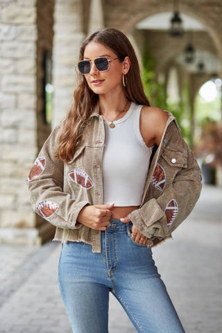 Woman wearing a beige jacket with football designs, white top, and blue jeans outdoors.