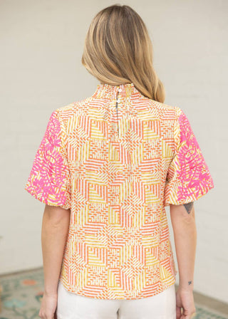 A woman viewed from the back wearing a short-sleeve collared blouse with a geometric pattern in yellow, orange, and pink, paired with white shorts.