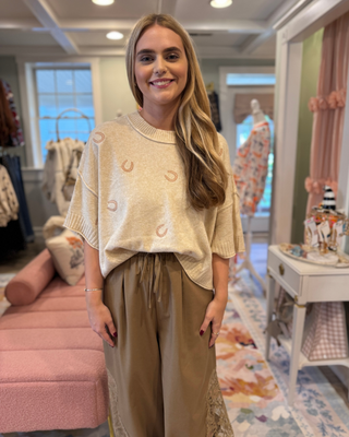 Woman wearing an oatmeal oversized short sleeve knit sweater with blush pink horseshoe accents, ribbed neckline, and cuffs, styled with taupe pants.