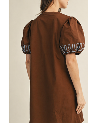 Women's saddle brown shift dress with white embroidered trim, puff sleeves, and a relaxed silhouette, styled with ivory cowboy boots for a western-inspired fall boutique outfit.
