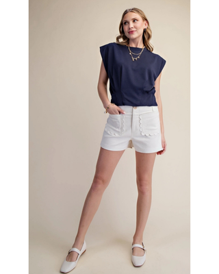 Woman modeling the white Emerson High-Waisted Shorts with scalloped patch pockets and a navy sleeveless top.