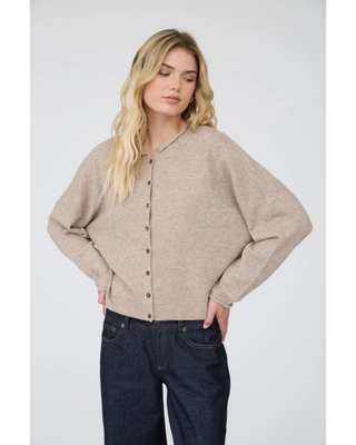 A blonde woman wearing a light beige knit cardigan with a buttoned front and dark blue jeans, photographed against a neutral light gray studio background.