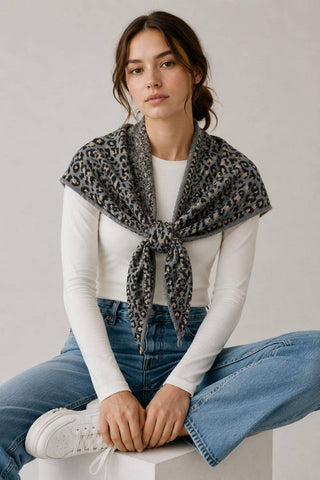 A woman wearing a leopard print scarf tied around her neck over a white long-sleeve top and blue jeans, sitting on a white block against a light gray background.