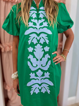 Woman wearing an emerald green embroidered shift dress with white appliqué detailing and puff sleeves, styled for a chic boutique fashion look.