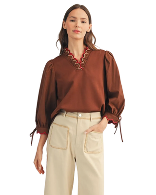 Women’s chocolate brown blouse with red scalloped trim, decorative V-neckline, puff sleeves and tie cuffs, styled with cream pants.